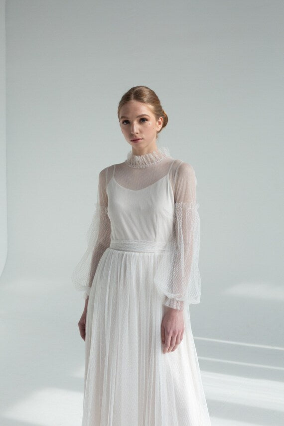 A woman standing in a white tulle wedding dress with floral sleeves and a satin top, against a white background.