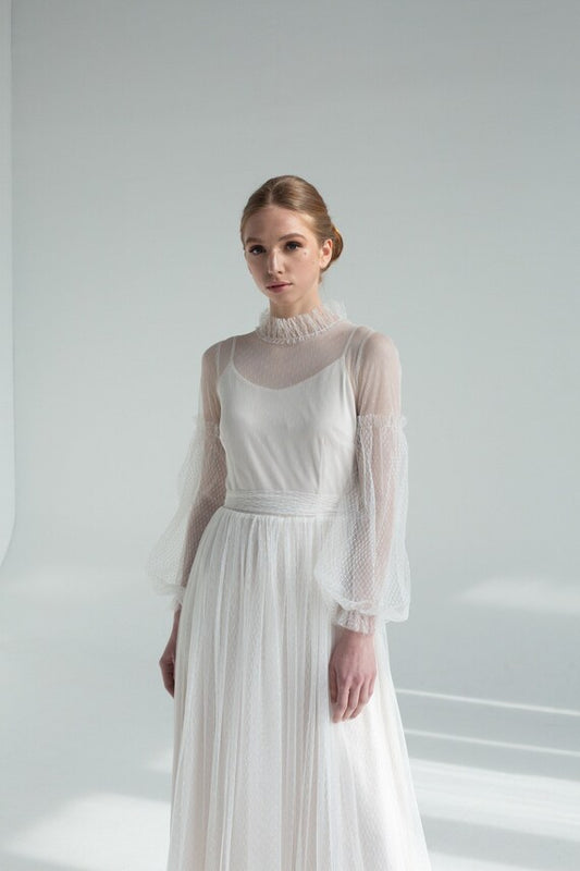 A woman standing in a white tulle wedding dress with floral sleeves and a satin top, against a white background.