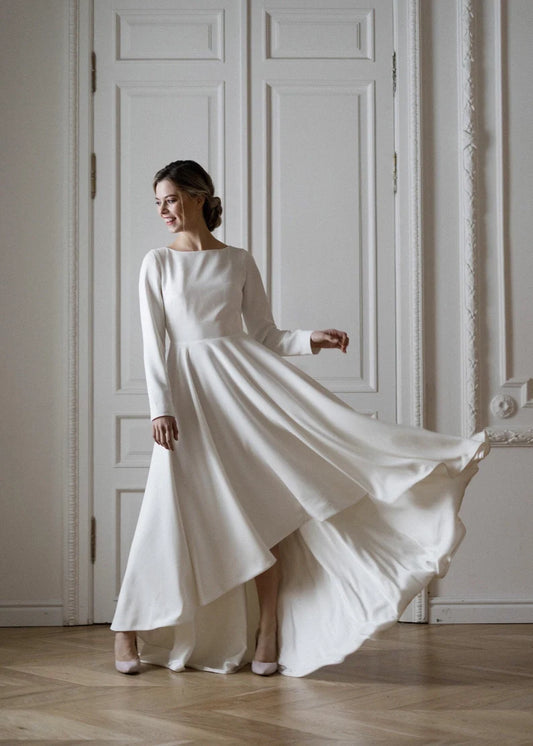 A woman in a white, high-low skirt wedding dress with short sleeves, standing in a room with wooden flooring and a door in the background.