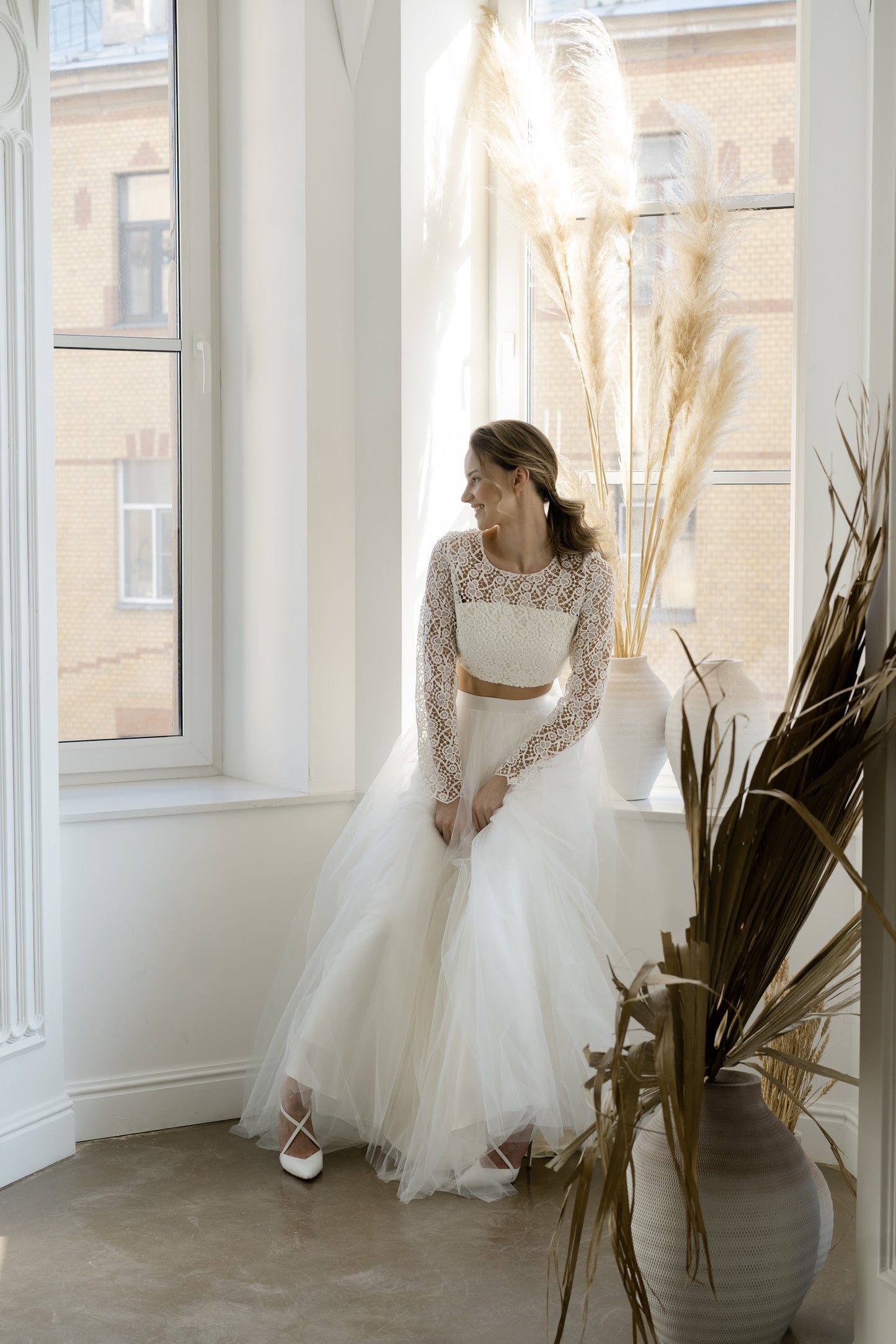 Bride in Margaux bridal two-piece sitting by a window, full length