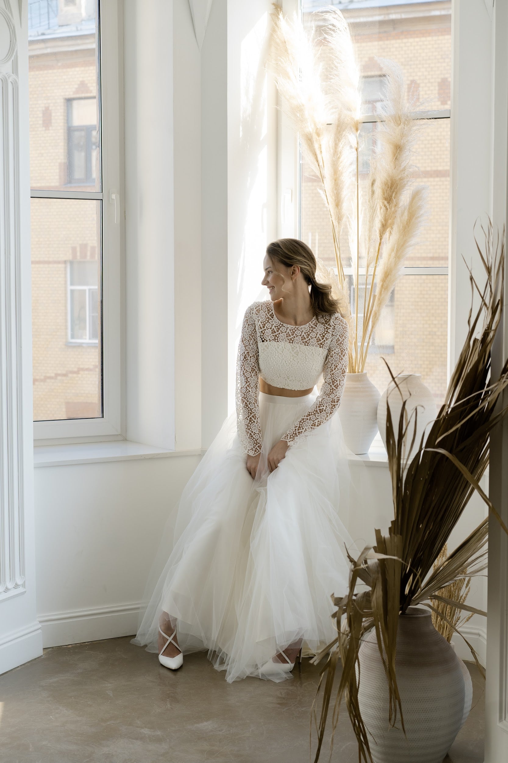 Bride in Margaux bridal two-piece sitting by a window, full length